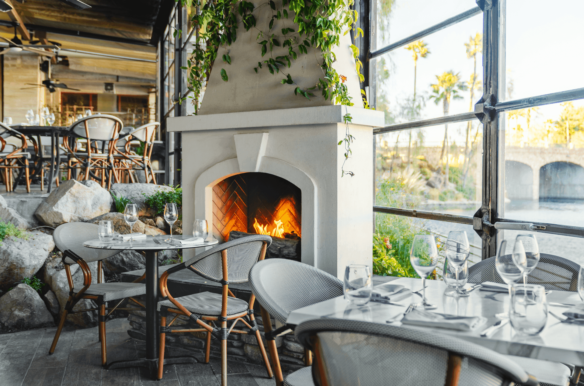 Cozy restaurant interior with fireplace and set dining table near large window overlooking outdoor scene.